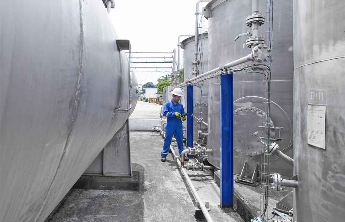 Person in blue coveralls, hard hat, and yellow gloves checking tablet standing in between large tanks