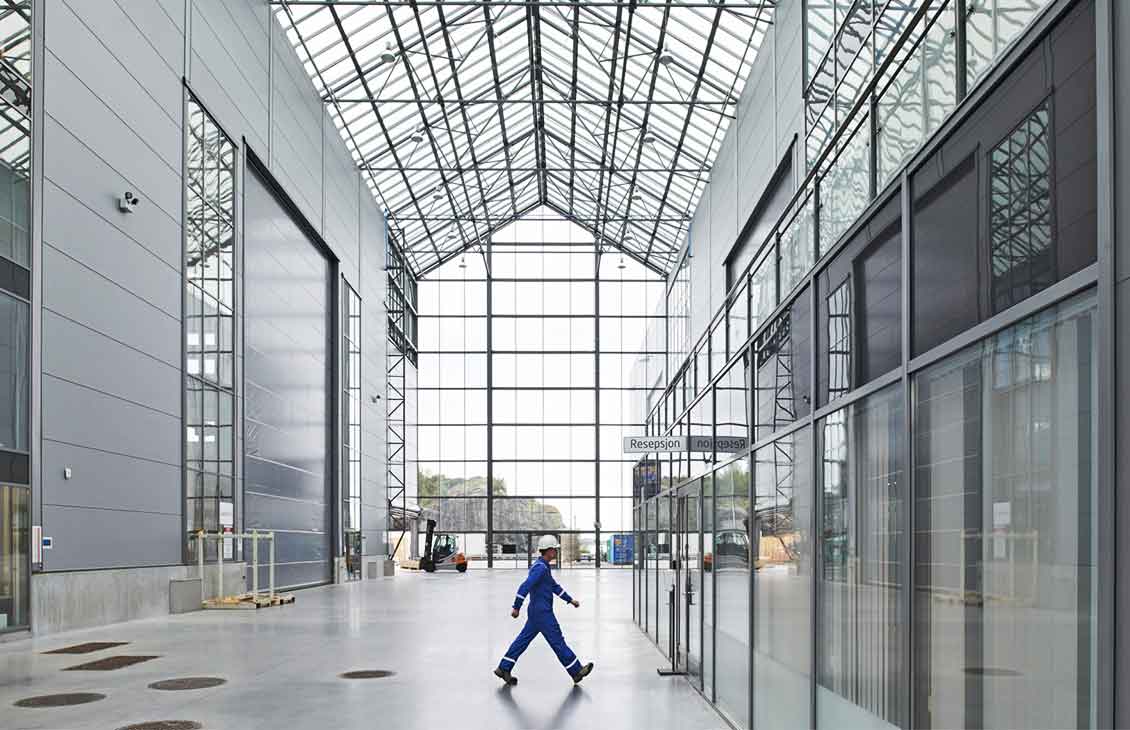 Man in blue coveralls and white hardhat walking in warehouse with glass roof